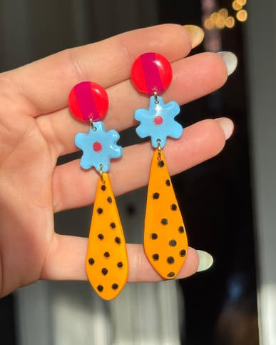 Colorful earrings with red, blue, and orange designs held in a hand against a blurred background.